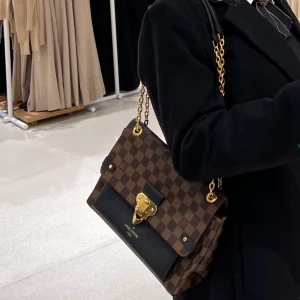 Louis Vuitton väska - Snygg brun rutig handväska från Louis Vuitton med gulddetaljer och kedjehandtag. Väskan har en elegant låsdetalj framtill och är perfekt för att ge en lyxig touch till din outfit. Köptes för 23 000kr, kvitto finns. Helt i nyskick! 