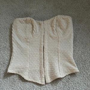 Beige broderad korsettopp från Divided - Säljer en beige korsettopp från Divided i storlek XXS. Toppen är axelbandslös och har ett broderat mönster över hela tyget. Perfekt att styla till olika outfits för en trendig look.