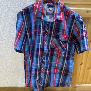 Rutig kortärmad skjorta i blått och rött - Säljer en färgglad kortärmad skjorta från Columbia med rutigt mönster i blått, rött och vitt. Skjortan har klassisk krage, bröstficka med knapp och knäppning framtill. Perfekt för dig som gillar en avslappnad och stilren look.