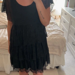 Svart volangklänning med spetsdetaljer - Säljer en svart kortklänning med flera lager volanger och fina spetsdetaljer. Klänningen har korta, transparenta ärmar i spets och rund halsringning. Perfekt för dig som gillar romantisk och feminin stil.