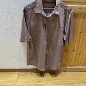 Brun kortärmad skjorta med diskret mönster från Selected Homme - Säljer en brun kortärmad skjorta från Sean John med diskret småmönster. Skjortan har klassisk krage, knappar framtill och axelremmar med knappdetalj. Perfekt för en avslappnad men stilren look.