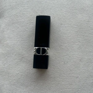 Rouge Dior läppstift 400 Nude Line Velvet - Säljer ett lyxigt Rouge Dior läppstift i färgen 400 Nude Line Velvet. Hylsan är svart med silverdetaljer och har den ikoniska CD-loggan. Läppstiftet har en djup, mörk nude-nyans med sammetsmatt finish.  Kommer utan förpackning, oanvänd och otestad, om du köper fler av dessa läppstift kan jag tänka mig bättre pris. Rensning inför flytt. Vissa märken på själva skalet på läppstiftet kan förekomma då jag förvarat alla i samma låda 