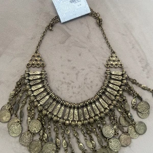 Guldigt statement-halsband med mynt - Unikt halsband i guldton med dekorativa hängande mynt och detaljerade mönster. Kedjan är grov och halsbandet har en bohemisk känsla med många rörliga delar. Perfekt för att lyfta en outfit med en touch av vintage.