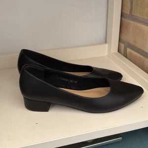 Svarta pumps med låg klack och spetsig tå - Stilrena svarta pumps med låg klack och spetsig tå. Skorna har en klassisk design och är tillverkade i ett slätt material med en mjuk innersula. Perfekta för dig som vill ha en elegant look.