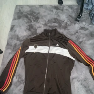 Brun Adidas track jacket med ränder - Säljer en brun Adidas track jacket med vit panel och texten 'The Brand' på ryggen. Jackan har dragkedja, höga muddar och klassiska Adidas-ränder i rött, orange och gult längs ärmarna. Perfekt för en sportig och retro look. Den har två fläckar där bak som går bort efter tvätten.
