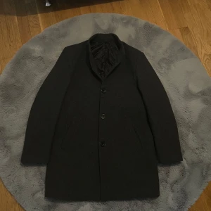 Mörkgrå rock med knappar - Mörkgrå rock med tre knappar framtill och diskret krage. Ytter- och innermaterialet är väldigt skönt och i bra kvalitet. Perfekt för kyligare dagar och ger en klassisk look.