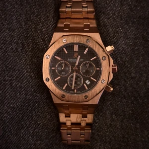 AP Klocks  - Exklusiv herrklocka från Audemars Piguet Royal Oak med guldigt armband och boett. Klockan har svart urtavla med kronograf och datumvisning. Ikonisk åttakantig form och detaljerad länkdesign.