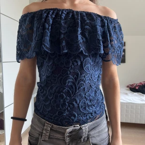 Volang body - Säljer en mörkblå offshoulder body i spets med vackert mönster. Blusen har korta ärmar och en volang upptill som ger en romantisk känsla. Perfekt för dig som vill ha en feminin och trendig topp.