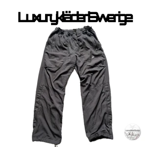 Nike byxor - Säljer ett vintage par grå cargobyxor från Nike med elastisk midja och dragsko vid benslut. Storlek M-L. Byxorna har flera fickor och en broderad Nike-logga på benet. Perfekta för en avslappnad och sportig stil.