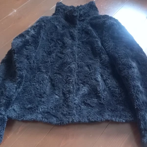 Svart teddyjacka med hög krage - Mysig svart teddyjacka med hög krage och knappar framtill. Jackan har lång ärm och är fodrad på insidan för extra komfort. Perfekt för kyliga dagar när du vill hålla dig varm och snygg.