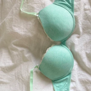Mintgrön bh från H&M med spetsdetaljer - Säljer en mintgrön bh från H&M med vadderade kupor och fina vita spetsdetaljer längs kanterna. Bh:n har en liten rosett framtill och justerbara axelband. Tillverkad i bomullsmix för extra komfort.