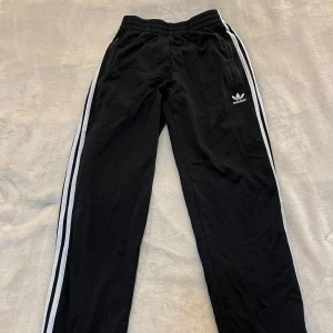 Svarta Adidas byxor med vita ränder - Svarta Adidas byxor med klassiska vita ränder längs sidorna och Adidas-logga på ena benet. Byxorna har resår i midjan och är tillverkade i ett syntetmaterial som passar perfekt till träning eller en sportig stil.