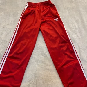 Röda Adidas byxor med vita ränder - Säljer ett par klassiska röda Adidas byxor med vita ränder längs sidorna och Adidas-logga på låret. Byxorna har resår i midjan och är raka i modellen, perfekta för en sportig och avslappnad stil.