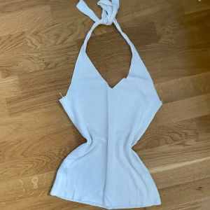 Vit ribbad halterneck linnetopp - Säljer en vit ribbad halterneck topp med knyt i nacken. Toppen har en figurnära passform och är ärmlös, perfekt för varma dagar eller som ett snyggt lager under en jacka.