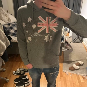 Grå hoodie med australienskt flaggtryck - Säljer en grå hoodie med tryck av Australiens flagga i rött och vitt på bröstet. Tröjan har huva med dragsko och en stor magficka. Perfekt för dig som gillar en avslappnad stil. Jag är 185cm. Tröjan är storlek M.