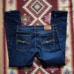 Mörkblå jeans från Nudie Jeans - Snygga blå jeans från Nudie Jeans med klassisk femficksdesign och kontrastsömmar. De har en slim passform och är så kallade dry/raw och därmed har de en unik och väldigt snygg tvättad look, inga är alltså likadana. Stå ut från mängden med dessa enastående jeans, 1 av 1 par. Storlek W29, sitter som 30, L32. 