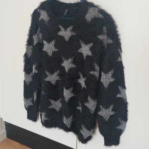 Svart fluffig tröja med stjärnmönster från Laura Torelli Collection - Säljer en svart fluffig tröja från Laura Torelli Collection i storlek M. Tröjan har ett tydligt stjärnmönster i grått och är långärmad. Perfekt för dig som gillar unika och mysiga plagg med lite extra attityd.