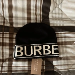 Svart mössa från Burberry  - Svart mössa från Burberry med bred vit kant och stor stickad logotyp runt om. Klassisk och stilren design med tydlig märkning. Perfekt accessoar för kyligare dagar.