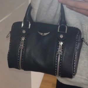 Svart handväska med silverdetaljer, Sunny XS studs bag  - Snygg svart handväska i skinnimitation med två dekorativa dragkedjor framtill och silverfärgade nitar längs kanterna. Väskan har dubbla handtag och ett emblem i silver på framsidan. Perfekt för att lyfta din outfit. Lite använd och mycket bekväm, har även bandet för att använda som Cross body också. Priset kan diskuteras. Har även tyväskan som jag jag fick den i när jag köpte den. 