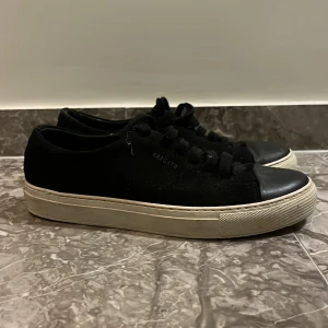 Svarta sneakers från Axel Arigato - Svarta sneakers från Axel Arigato med vit platt sula och rund tå. Skorna har snörning och är tillverkade i skinn. Perfekta för en stilren och enkel look.