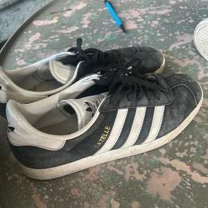 Adidas Gazelle sneakers i svart och vitt - Säljer ett par klassiska Adidas Gazelle sneakers i svart mocka med vita detaljer och de ikoniska tre ränderna på sidorna. Skorna har snörning och vit platt sula. Perfekta för en stilren och sportig look.