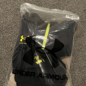 Svart träningströja från Under Armour - Svart träningströja från Under Armour med gul logga på bröstet och dragkedja framtill. Perfekt för sport och träning, tillverkad i syntetmaterial för bra andningsförmåga. Tröjan har lång ärm och sportig design.
