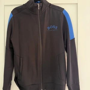 Svart track jacket från Hugo Boss med blå detaljer - Svart track jacket från Hugo Boss med blå logga på bröstet och blå paneler på axlarna. Jackan har dragkedja framtill, ståkrage och två fickor. Perfekt för en sportig och stilren look. Använd max 5 gånger så väldigt bra skick.