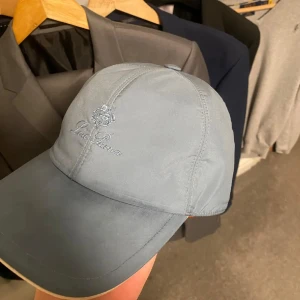 Blå keps från Loro Piana - Snygg blå keps från Loro Piana med broderad logotyp framtill. Perfekt för en stilren look.  Kepsen har lite fläckar framme vid där skärmen börjar samt under skärmen och ovanför spännet men inget som man tänker på särskilt mycket men därav priset. Vid fler bilder är det bara att fråga!