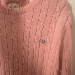 Rosa kabelstickad tröja från GANT - Säljer en klassisk rosa kabelstickad tröja från GANT med rund halsringning och broderad logga på bröstet. Tröjan har långa ärmar använd 2 gnr