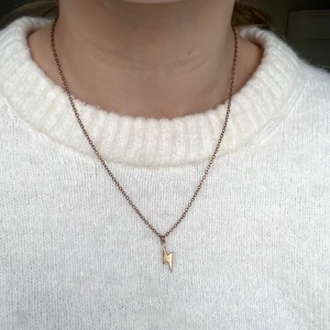 Halsband med blixtberlock i guld - Säljer ett stilrent halsband med tunn kedja i guld och en blixtformad berlock. Perfekt för dig som gillar minimalistiska smycken med en cool detalj.