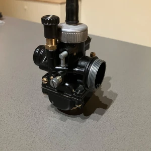 Svart förgasare till moped eller motorcykel - Säljer en svart förgasare i metall med detaljer i silver och mässing. Passar till moped eller motorcykel. Kompakt design med flera justerskruvar och anslutningar för bränsle och luft.