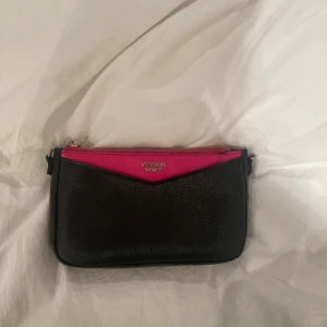 Victoria's Secret axelväska  - Säljer en snygg svart axelväska från Victoria's Secret med en rosa detalj upptill och silverfärgad dragkedja. Väskan har en justerbar axelrem med kedjedetaljer och är perfekt för att bära det viktigaste. Loggan syns framtill på väskan. Vet inte bara om den är riktig eller inte men jag tror inte det tyvärr✨