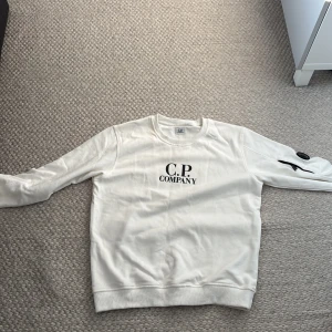 Vit sweatshirt från C.P. Company - Säljer en vit sweatshirt från C.P. Company med svart logga på bröstet och en ikonisk linsdetalj på ärmen. Tröjan har rund halsringning och ribbade muddar. Perfekt för en stilren och modern look. Använd bara en gång och är i jättebra skick.
