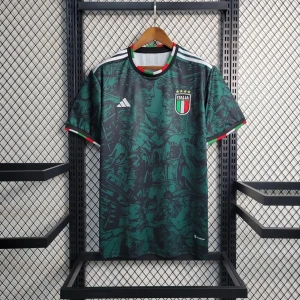 Italien fotbollströja Adidas - Snygg grön fotbollströja från Adidas med Italiens landslagsmärke på bröstet. Tröjan har korta ärmar, vita Adidas-ränder på axlarna och röda detaljer vid krage och ärmslut. Mönstret är mörkgrönt med svarta inslag.