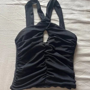 Svart halterneck topp från Gina Tricot - Säljer en svart halterneck topp från Gina Tricot med snygg rynkning framtill och en liten öppning vid bysten. Toppen har breda band som knyts i nacken och är figurnära i modellen. Perfekt för att lyfta din outfit!