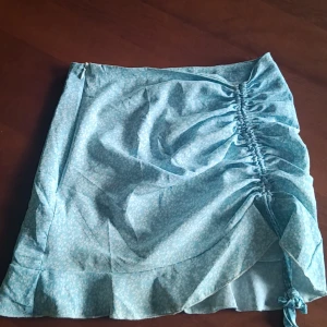 Blommig kortkjol från Shein - Söt blommig kortkjol från Shein i ljusblått med vita blommor. Kjolen har en volangdetalj och en knytning på sidan för en justerbar passform. Perfekt för varma dagar! Lite smutsig längst ner