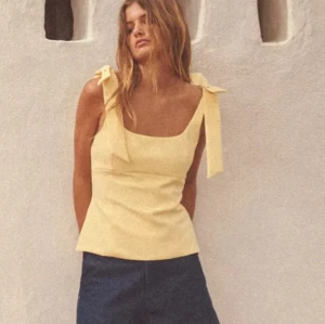 Gul linnetopp med rosetter på axlarna - Säljer en söt ljusgul linnetopp med breda axelband som knyts i rosetter. Toppen har en fyrkantig urringning och är figursydd för en feminin siluett. Perfekt för varma dagar och enkel att styla med både kjol och jeans.