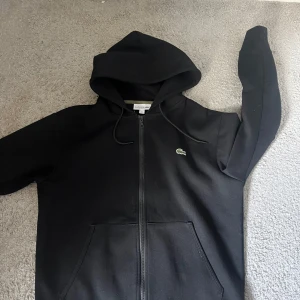 Lacoste hoodie - Säljer en svart hoodie från Lacoste med dragkedja och klassisk krokodillogga på bröstet. Tröjan har huva med snörning och två fickor framtill. Perfekt 10/10 skick hör av er vid minsta fundering