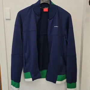 Blå träningsjacka från Björn Borg - Sportig blå träningsjacka med gröna och vita muddar från Björn Borg. Jackan har dragkedja framtill, lång ärm och en stilren design med liten logotyp på bröstet. Perfekt för träning eller en aktiv vardag.