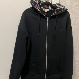  Burberry tröja - Säljer en svart hoodie från Burberry med hel dragkedja och klassiskt rutigt foder i huvan. Tröjan har två fickor framtill och justerbar huva med snören. Perfekt för en stilren och bekväm look. helt äkta