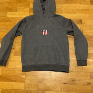 Grå hoodie från GANT  - Säljer en grå hoodie från GANT med broderad logga och ett sköldmärke med stjärnor och ränder på bröstet. Tröjan har huva och en stor magficka framtill. Perfekt för en avslappnad stil.