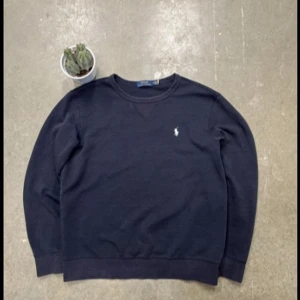 Mörkblå sweatshirt från Polo Ralph Lauren - Säljer en klassisk mörkblå sweatshirt från Polo Ralph Lauren med rund halsringning och broderad vit logga på bröstet. Tröjan har långa ärmar och är tillverkad i mjuk bomull. Perfekt för en stilren och avslappnad look.