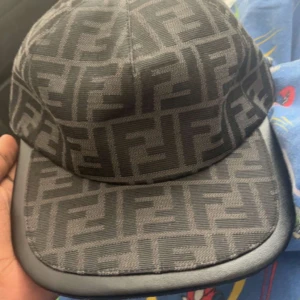 Svart och grå keps från Fendi - Säljer en snygg keps från Fendi i svart och grått med det klassiska FF-mönstret över hela kepsen. Kepsen har böjd skärm med läderdetalj och är tillverkad i ett slitstarkt tyg. Perfekt för dig som gillar exklusiva accessoarer.