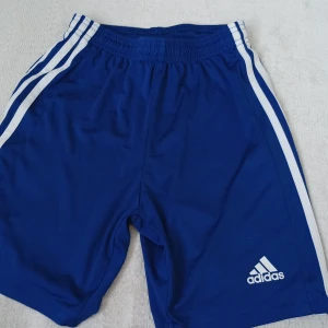 Blå träningsshorts från Adidas - Snygga blå träningsshorts från Adidas med vita ränder längs sidorna och logga framtill. Modellen har elastisk midja och är tillverkad i lätt och funktionellt material med AEROREADY-teknologi.