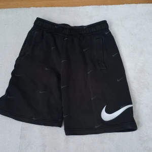 Svarta shorts från Nike med vit logga - Svarta shorts från Nike med elastisk midja och snörning. Shortsen har flera små Nike-loggor i grått och en stor vit Swoosh på ena benet. Två sidofickor. Perfekta för träning eller chill.