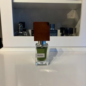 Nasomatto pardon - Unik parfym med grön vätska i en fyrkantig glasflaska och ett distinkt brunt trälock. Märkt med 'Pardon' på framsidan. Flaskan har en exklusiv och modern design som sticker ut i parfymhyllan.