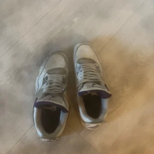 Vita sneakers Nike Air Jordans med lila detaljer - Säljer ett par vita Nike Air Jordans sneakers med lila detaljer. Skorna har snörning, meshpaneler och en platt sula. Klassisk design med rund tå och synlig Air-dämpning i sulan. Perfekta för dig som gillar streetwear och vill sticka ut med en ikonisk modell.