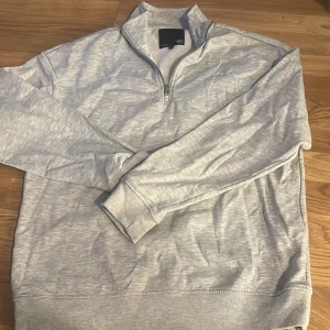Ljusgrå halvzip tröja från 157 - Säljer en ljusgrå tröja från 157 med halv dragkedja och hög krage. Tröjan har långa ärmar och ribbade muddar vid ärmslut och nederkant. Perfekt för lager-på-lager och enkel att matcha med det mesta.