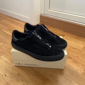 Axel arigato skor - Säljer ett par svarta sneakers från Axel Arigato i mocka med dragkedja på ovansidan istället för klassisk snörning. Skorna har en låg profil och platt sula, perfekt för en stilren look. Dom är också även mycket unika då dom inte längre säljs nya. Priset är diskuterbart.