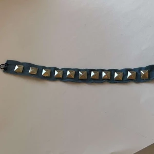 Blått armband med nitar - Säljer ett blått tygarmband med silverfärgade fyrkantiga nitar. Armbandet har ett svart spänne för enkel stängning. Perfekt för dig som gillar stil.